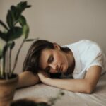 Woman sleeping with her head and arms laying on desk, next to house plant.